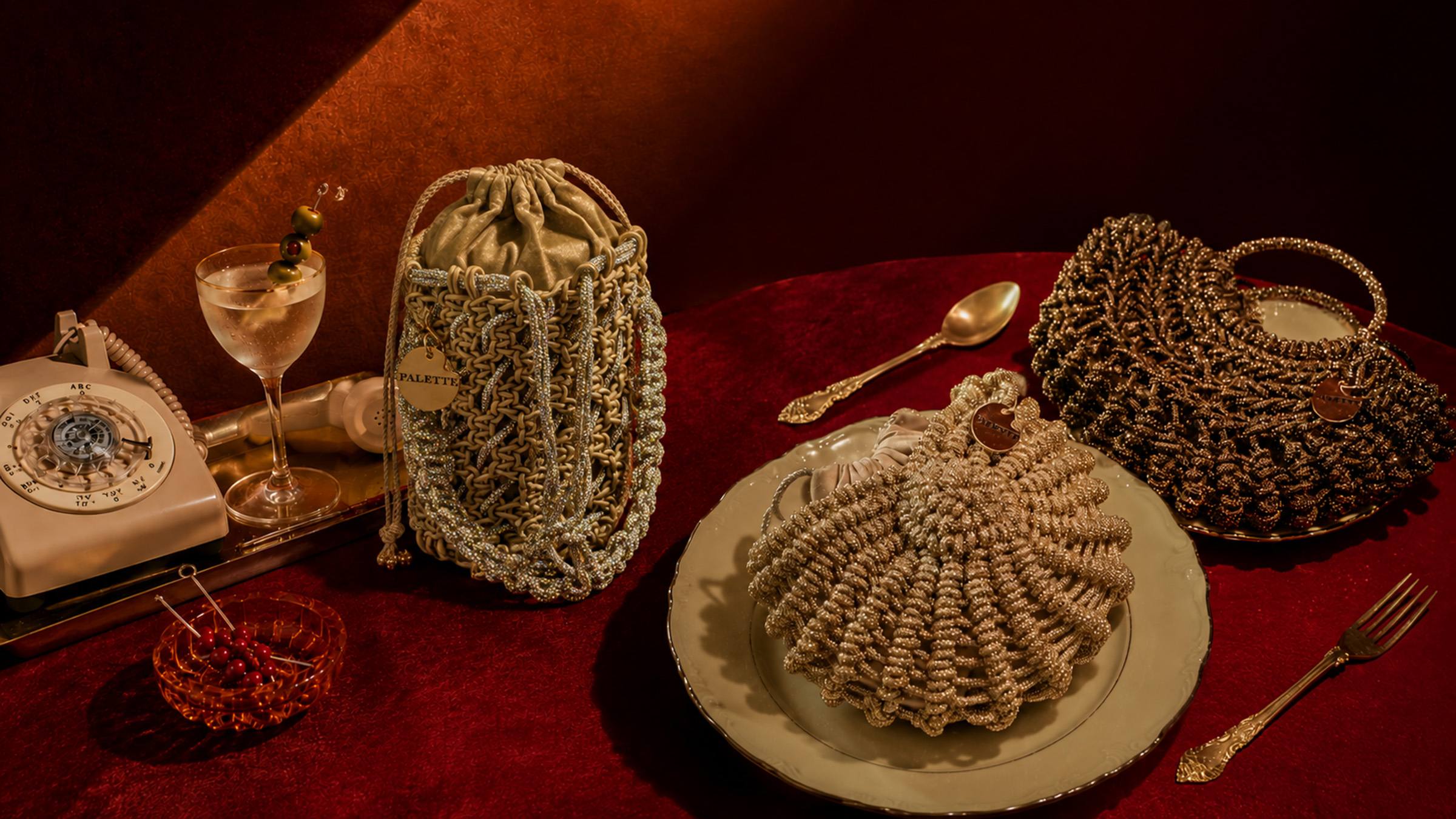 Three Palette by Rishma handcrafted bags — an ivory-gold drawstring bucket, a champagne pouf, and an onyx tote — arranged on a deep red velvet table alongside a martini glass, vintage rotary phone, and ornate silver cutlery, in a warm amber-lit setting.