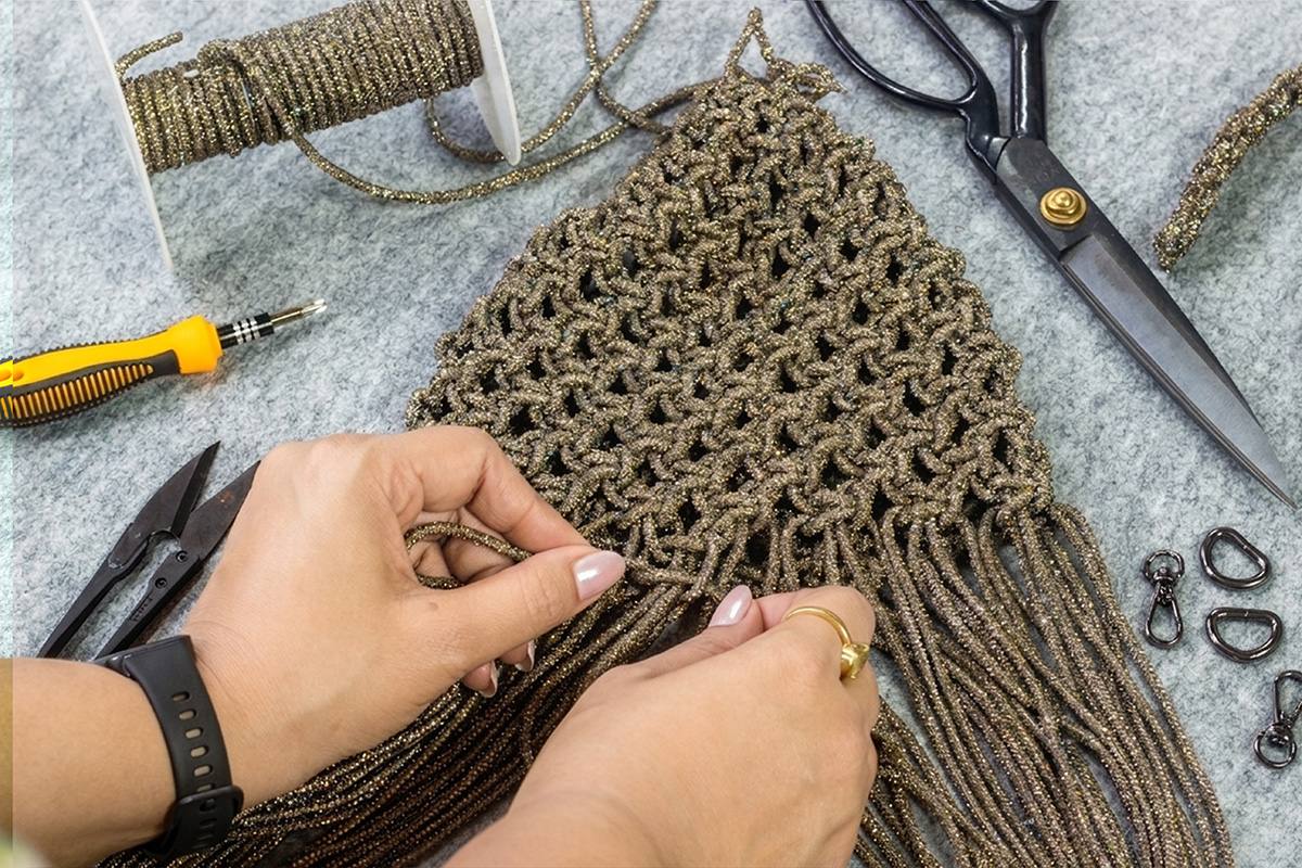 Person working on a macrame bag with tools on a gray surface - Palette by Rishma